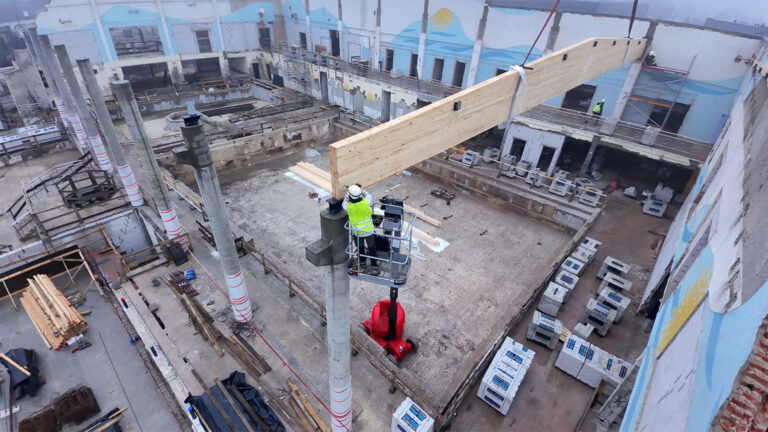 Perspektywa z drona pokazująca pracownika w podnośniku koszowym mocującego drewniany wspornik dachu.