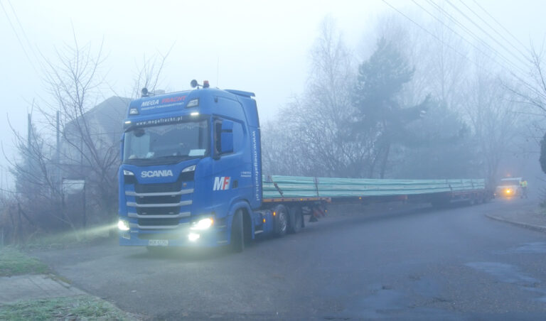 Ciągnik siodłowy w asyście pilota wiozący elementy konstrukcji dachu.